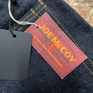 May include: Close-up of dark blue denim fabric with yellow stitching. A red tag with the text "JOE McCOY" and "REAL WORK CLOTHING" is attached. The tag also includes the text "THE CATTLE DRIVE BRAND".