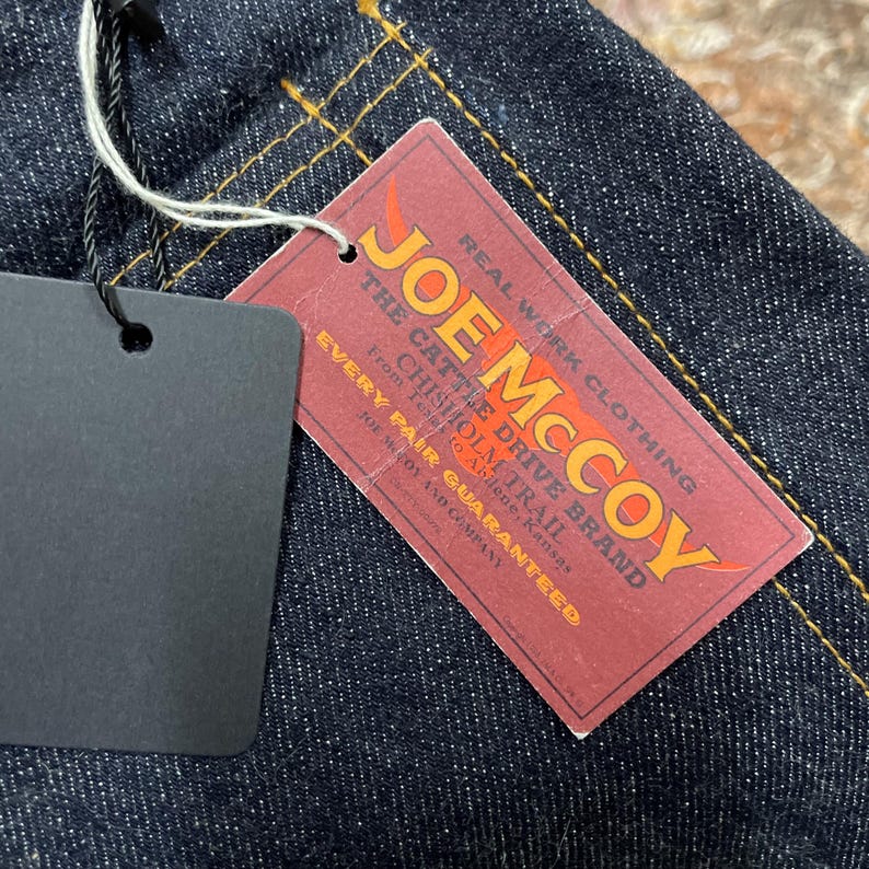 May include: Close-up of dark blue denim fabric with yellow stitching. A red tag with the text "JOE McCOY" and "REAL WORK CLOTHING" is attached. The tag also includes the text "THE CATTLE DRIVE BRAND".