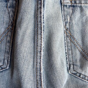 May include: Close-up of light blue denim fabric with visible stitching and pocket details. The denim has a faded appearance, with a focus on the texture and construction of the material. The stitching is in a contrasting brown colour.