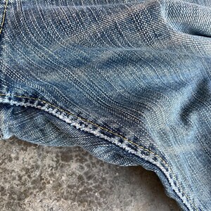 May include: Close-up of blue denim fabric with visible texture and stitching. The denim has a faded appearance, with lighter areas creating a worn look. Yellow stitching is prominent along the seams.
