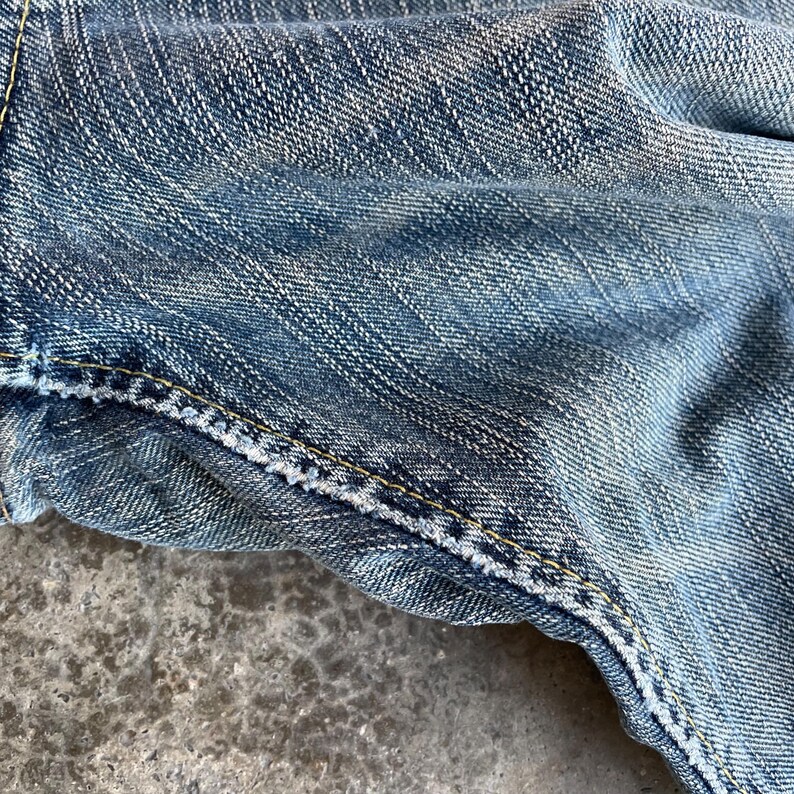 May include: Close-up of blue denim fabric with visible texture and stitching. The denim has a faded appearance, with lighter areas creating a worn look. Yellow stitching is prominent along the seams.