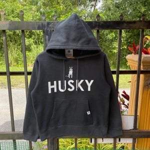 Puede incluir: Sudadera con capucha gris oscuro con bolsillo delantero y capucha. La palabra "HUSKY" está impresa en letras blancas grandes en el pecho, con un gráfico de un perro husky encima. La sudadera tiene un cordón y un pequeño logotipo en la capucha.
