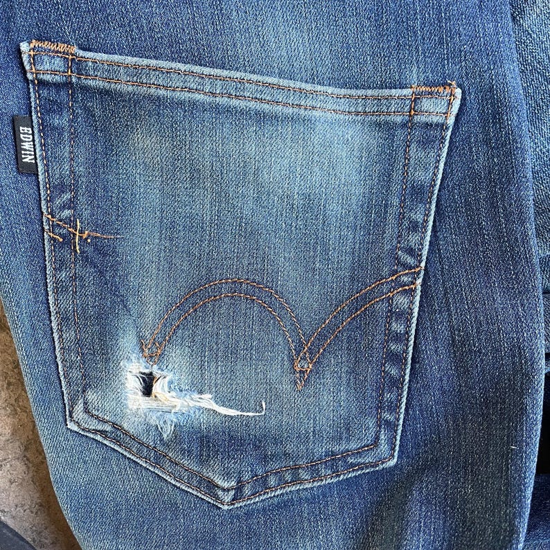 Op de afbeelding: Close-up van een blauwe denim jeans. De jeans heeft een beschadigde zak met een scheur en zichtbare stiksels. Het Edwin-merklabel is zichtbaar. De denim heeft een vervaagde, versleten uitstraling.