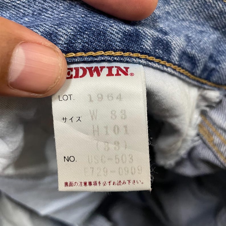 May include: A white fabric clothing tag sewn into blue denim jeans. The tag features the red text EDWIN, followed by LOT. 1964, size information W 83, H 101, and NO. U5C-503. Japanese text is printed at the bottom of the tag, and yellow stitching is visible.