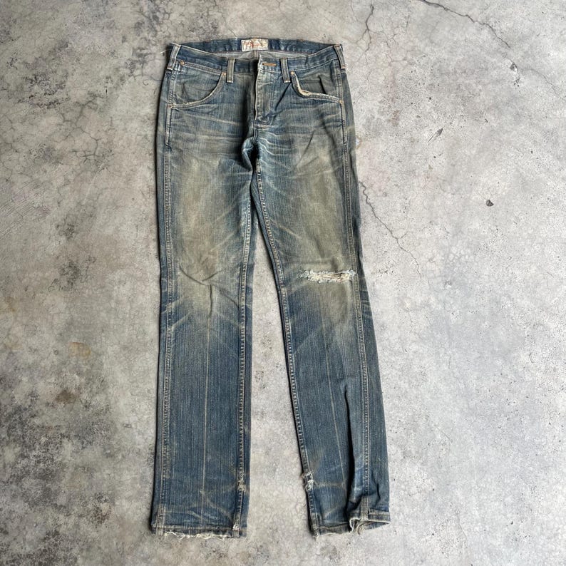 May include: Faded blue denim jeans with a distressed appearance. The jeans feature a button fly, classic five-pocket design, and a ripped knee. The denim has a worn, vintage look.
