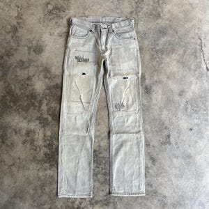 K&ouml;nnte beinhalten: Hellgraue Jeans aus Denim mit abgenutzten Patches und dekorativen N&auml;hten. Die Jeans haben ein klassisches F&uuml;nf-Taschen-Design und einen Knopf- und Rei&szlig;verschluss. Die Patches sind auf der Vorderseite der Jeans aufgen&auml;ht.