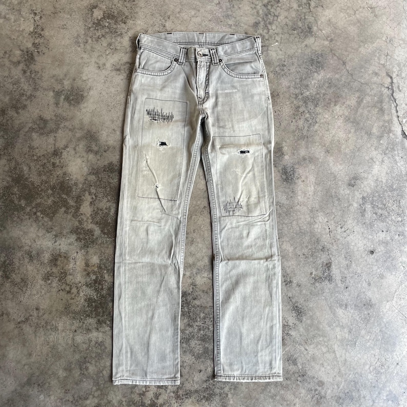 K&ouml;nnte beinhalten: Hellgraue Jeans aus Denim mit abgenutzten Patches und dekorativen N&auml;hten. Die Jeans haben ein klassisches F&uuml;nf-Taschen-Design und einen Knopf- und Rei&szlig;verschluss. Die Patches sind auf der Vorderseite der Jeans aufgen&auml;ht.