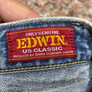 May include: A rectangular red fabric label stitched onto blue denim fabric. The label features yellow text reading ONLY GENUINE EDWIN US CLASSIC and white text reading PRODUCED BY EDWIN COMPANY LIMITED. The denim has visible yellow stitching.