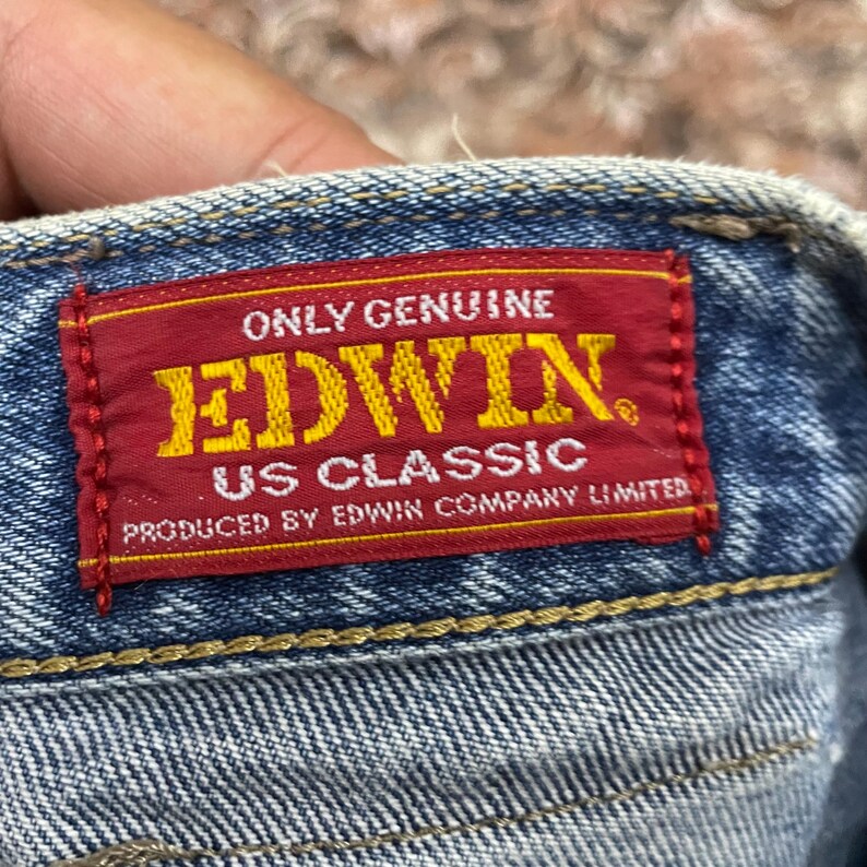 May include: A rectangular red fabric label stitched onto blue denim fabric. The label features yellow text reading ONLY GENUINE EDWIN US CLASSIC and white text reading PRODUCED BY EDWIN COMPANY LIMITED. The denim has visible yellow stitching.