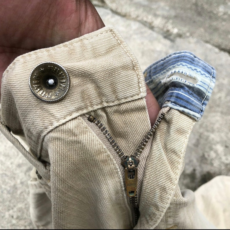 May include: Close-up of a pair of beige denim pants with a zipper and a metal button. The pants have a worn look and are made of a thick, durable fabric.