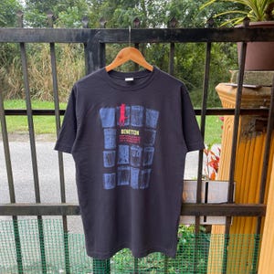May include: Black t-shirt with a graphic print of a cat and a grid of blue and white cans. The text "Benetton Are You Looking For Your Own Drink On A Monday And T" is printed on the shirt.