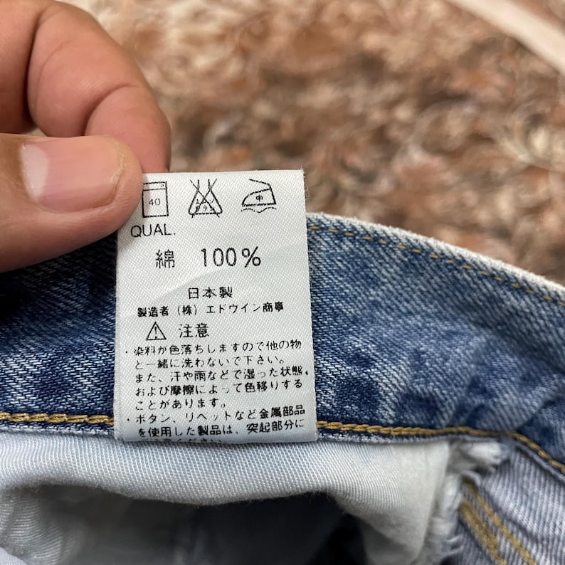 May include: A white fabric care label attached to blue denim jeans. The label features black text including '100%' and Japanese characters, along with three laundry instruction symbols at the top. The denim has visible yellow stitching.