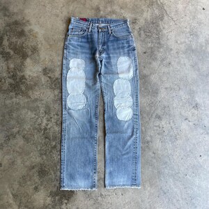 Könnte beinhalten: Hellblaue Jeans mit hellblauen Aufnähern auf der Vorderseite jedes Beins. Die Jeans haben ein klassisches Fünf-Taschen-Design und einen geraden Beinverlauf. Die Aufnäher sind oval geformt und in einem vertikalen Muster angeordnet.