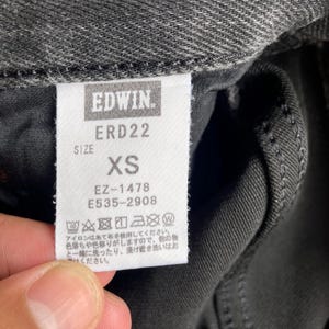 May include: Close-up of a tag from Edwin jeans. The tag is white with black text, displaying the brand name, style number ERD22, and size XS. Additional codes EZ-1478 and E535-2908 are also printed on the tag. The tag is attached to dark gray denim.