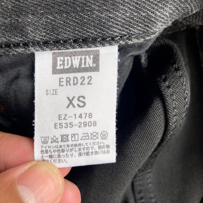 May include: Close-up of a tag from Edwin jeans. The tag is white with black text, displaying the brand name, style number ERD22, and size XS. Additional codes EZ-1478 and E535-2908 are also printed on the tag. The tag is attached to dark gray denim.