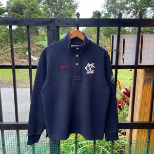 Op de afbeelding: Marineblauwe pullover met lange mouwen en een kraag met knopen. Het heeft een wit geborduurd logo op de borst en een klein rood logo aan de linkerkant. De trui heeft een relaxte pasvorm en is gemaakt van een zacht materiaal.