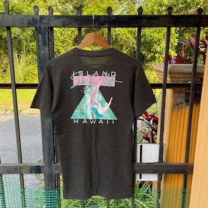 May include: Dark gray t-shirt with a graphic on the back featuring a surfer riding a wave. The design includes the words "ISLAND Surf Shop HAWAII" in a stylized font with teal and pink accents. The shirt is hanging on a wooden hanger.