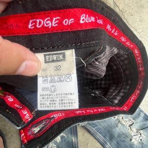 K&ouml;nnte beinhalten: Nahaufnahme der Innenseite einer blauen Jeans. Die Jeans hat einen schwarzen Bund mit roter Paspel, auf dem "EDGE OF BLUE" steht. Ein Etikett zeigt "EDWIN" und "SIZE 32". Die Jeans hat einen Used-Look.