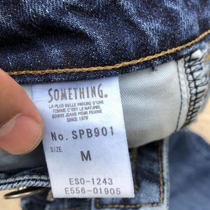 Op de afbeelding: Een blauw denim jeans-label met de merknaam "SOMETHING." en de tekst "LA PLUS BELLE PARURE D'UNE FEMME C'EST LE NATUREL EDWIN JEANS POUR FEMME SINCE 1979" en het modelnummer "SPB901". Het label bevat ook de maat "M" en de nummers "ESO-1243" en "E556-01905".