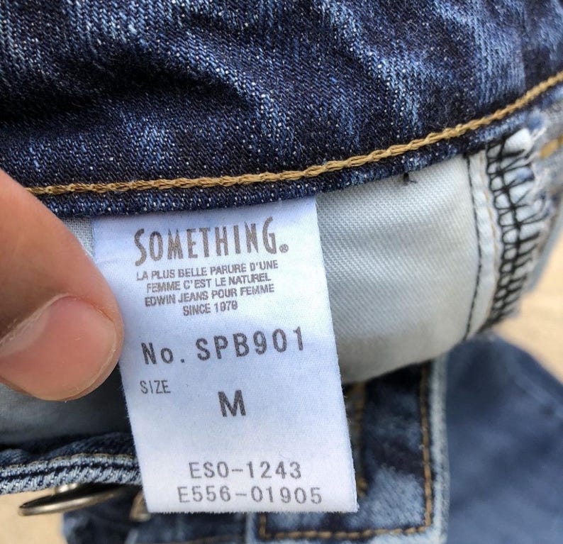 Op de afbeelding: Een blauw denim jeans-label met de merknaam "SOMETHING." en de tekst "LA PLUS BELLE PARURE D'UNE FEMME C'EST LE NATUREL EDWIN JEANS POUR FEMME SINCE 1979" en het modelnummer "SPB901". Het label bevat ook de maat "M" en de nummers "ESO-1243" en "E556-01905".