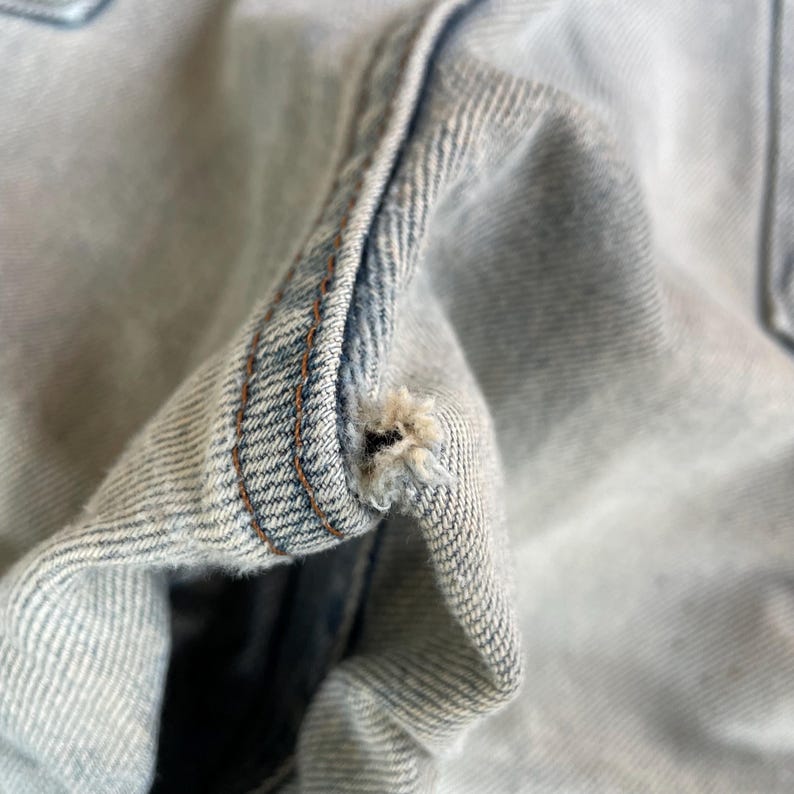 May include: Close-up of light blue denim fabric with a distressed, worn appearance. The image highlights a small tear and frayed edges, showcasing the texture and detail of the denim material. The stitching is visible.