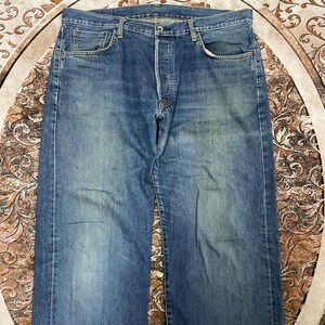 May include: A pair of blue denim jeans featuring a classic five-pocket design, a button fly closure, and a medium wash finish with subtle fading on the thighs. The jeans have a straight-leg cut, visible copper-toned rivets, and belt loops.