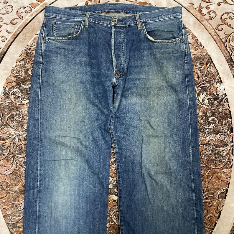 May include: A pair of blue denim jeans featuring a classic five-pocket design, a button fly closure, and a medium wash finish with subtle fading on the thighs. The jeans have a straight-leg cut, visible copper-toned rivets, and belt loops.