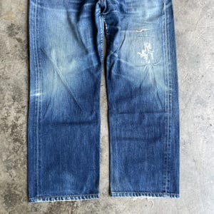 May include: Faded blue denim jeans with a straight leg cut. The jeans show signs of wear, including fading and a distressed patch on the right leg. The denim has a classic five-pocket design and a button and zip closure.