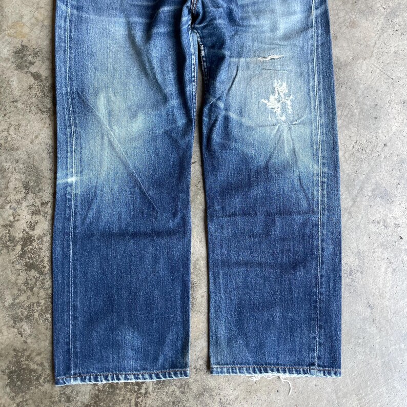 May include: Faded blue denim jeans with a straight leg cut. The jeans show signs of wear, including fading and a distressed patch on the right leg. The denim has a classic five-pocket design and a button and zip closure.