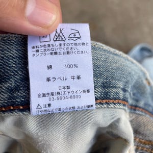 May include: A close-up of a white tag with Japanese text and care symbols, attached to blue denim fabric. The tag indicates the fabric is 100% cotton and has a leather label. The tag also includes the manufacturer's information.