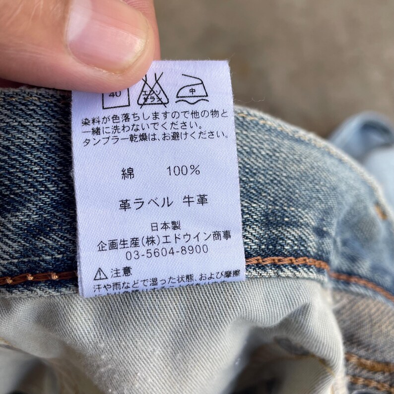 May include: A close-up of a white tag with Japanese text and care symbols, attached to blue denim fabric. The tag indicates the fabric is 100% cotton and has a leather label. The tag also includes the manufacturer's information.