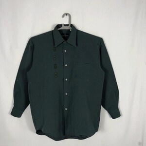 May include: A dark green long-sleeved button-down shirt with a small pocket on the left side. The shirt has a subtle pattern of small black squares on the left side.