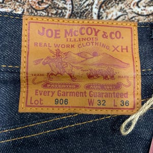 May include: Close-up of a brown leather label from Joe McCoy & Co. with text and a graphic of a cowboy on a horse. The label reads "Every Garment Guaranteed" and includes the size: W 32 L 36.