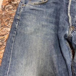 May include: A pair of blue denim jeans featuring a classic five-pocket construction with visible stitching, a button fly, and a faded wash effect across the front thigh area, showcasing the textured weave of the cotton fabric and metal rivets.