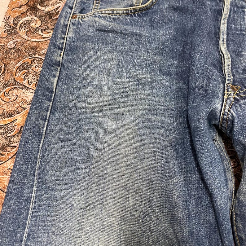 May include: A pair of blue denim jeans featuring a classic five-pocket construction with visible stitching, a button fly, and a faded wash effect across the front thigh area, showcasing the textured weave of the cotton fabric and metal rivets.