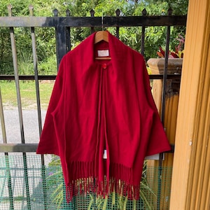 May include: A vibrant red cape with a large collar and a single button closure. The cape features a fringed hemline and is hanging on a wooden hanger. The garment is made of a soft material.