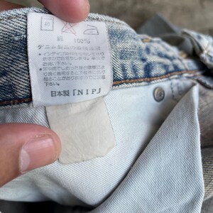 May include: Close-up of the inside of light-wash denim jeans. A white tag with Japanese text and care symbols is visible. The jeans are made of 100% cotton. The tag reads "Made in Japan [NIPJ]"