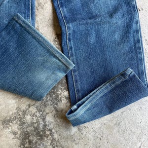 Op de afbeelding: Close-up van een blauwe spijkerbroek. De jeans heeft een klassiek ontwerp met vijf zakken, met een rechte pijp en een medium wassing. De stof is licht vervaagd, waardoor de jeans een vintage look krijgt. De zoom is opgevouwen.