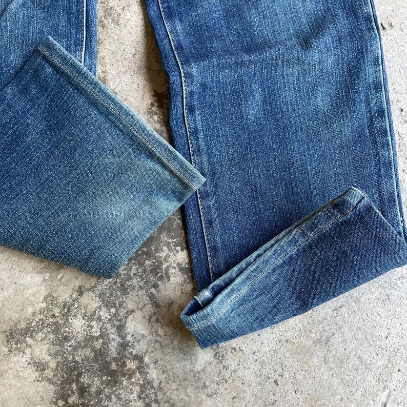 Op de afbeelding: Close-up van een blauwe spijkerbroek. De jeans heeft een klassiek ontwerp met vijf zakken, met een rechte pijp en een medium wassing. De stof is licht vervaagd, waardoor de jeans een vintage look krijgt. De zoom is opgevouwen.
