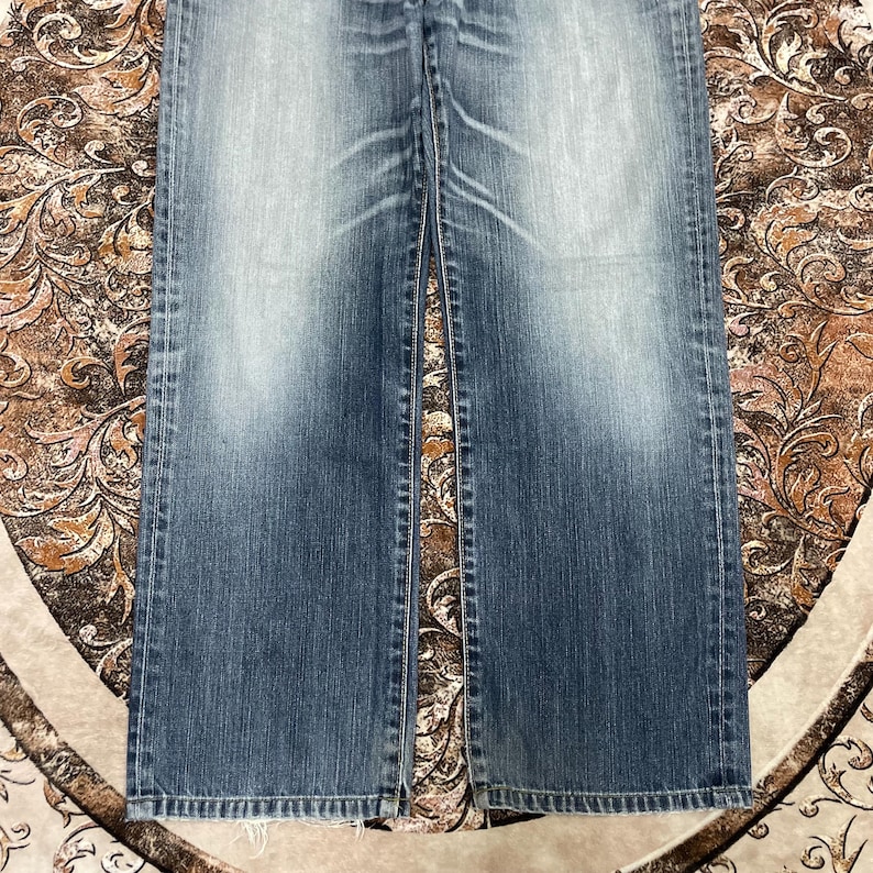 May include: A pair of blue denim jeans featuring a faded wash effect on the thighs and knees. The jeans have a straight-leg cut with visible stitching along the side seams and a classic five-pocket construction with a standard button fly closure.