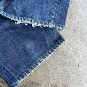 May include: Close-up of the bottom of blue denim jeans. The jeans have a frayed hem and are stitched with a contrasting thread. The fabric is a medium wash denim.