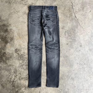 May include: Dark gray denim jeans with a faded wash and distressed details. The jeans feature classic five-pocket styling, a button and zipper closure, and a small logo patch on the back pocket. The jeans are laid flat on a concrete surface.