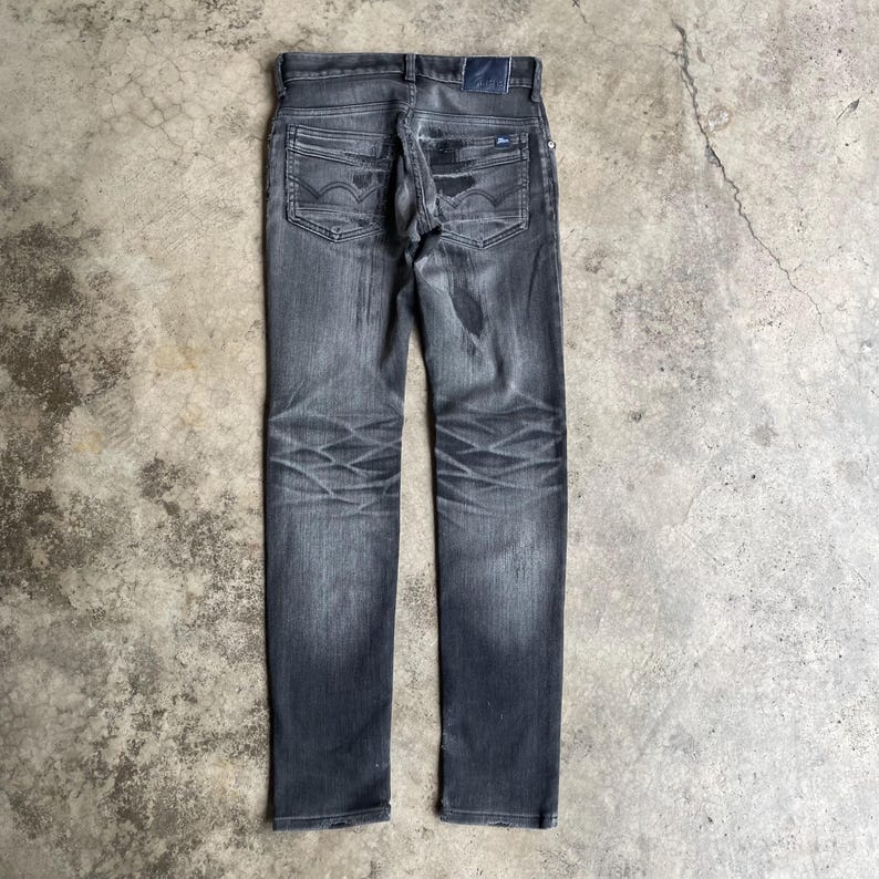 May include: Dark gray denim jeans with a faded wash and distressed details. The jeans feature classic five-pocket styling, a button and zipper closure, and a small logo patch on the back pocket. The jeans are laid flat on a concrete surface.