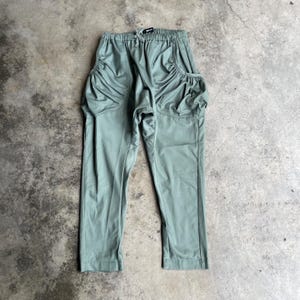 Op de afbeelding: Saliegroene broek met unieke, geplooide zakdetails. De broek heeft een tailleband met trekkoord en een relaxte pasvorm. De stof lijkt getextureerd. De broek ligt plat op een betonnen ondergrond.