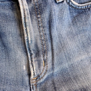 Pu&ograve; includere: Primo piano di jeans di denim blu, che mostra la patta, la tasca e i dettagli delle cuciture. Il denim ha un aspetto sbiadito con usura visibile, evidenziando la trama e la costruzione del capo.
