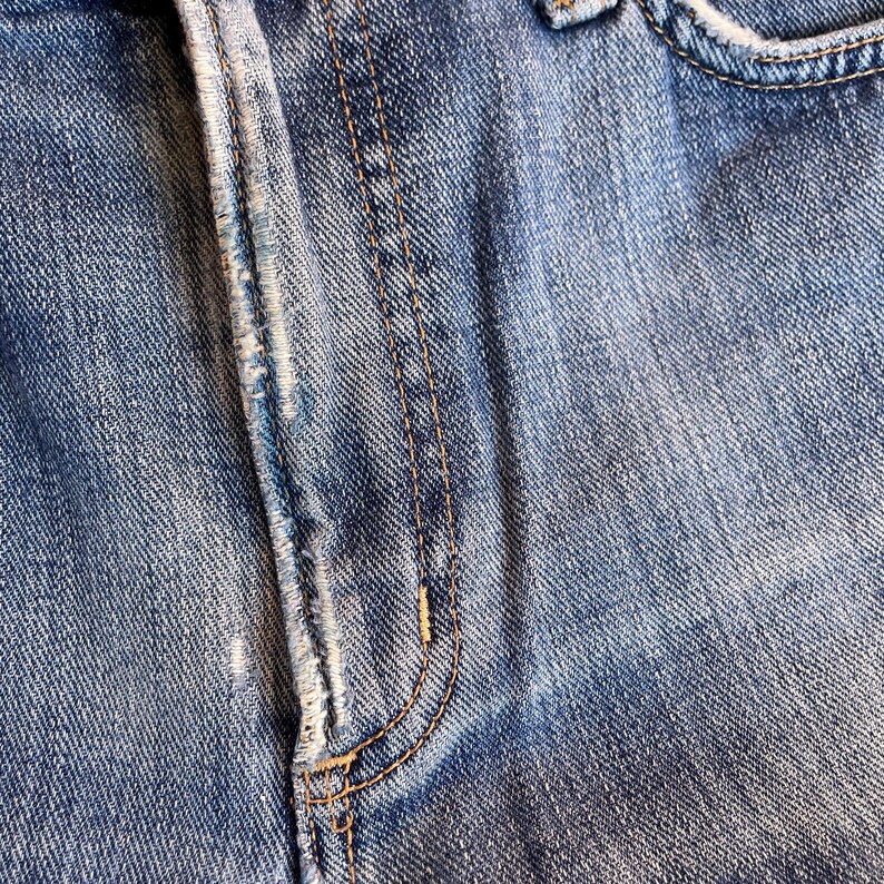 Pu&ograve; includere: Primo piano di jeans di denim blu, che mostra la patta, la tasca e i dettagli delle cuciture. Il denim ha un aspetto sbiadito con usura visibile, evidenziando la trama e la costruzione del capo.