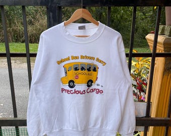 以下が含まれることがあります： 黄色いスクールバスのグラフィックと「School Bus Drivers Carry Precious Cargo」というテキストがプリントされた白いスウェットシャツ。