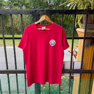 Könnte beinhalten: Ein rotes T-Shirt mit einem kleinen kreisförmigen Logo auf der oberen linken Brust. Das Logo zeigt den Text "Bubba Gump" in einem kreisförmigen Design. Das T-Shirt hängt an einem Holzbügel vor dem Hintergrund eines schwarzen Metallzauns und Grün.