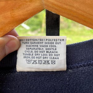 May include: A close-up of a navy blue garment tag with white text. The tag reads: 50% cotton/50% polyester, turn garment inside out, machine wash cool, separately, gentle cycle, do not bleach, tumble dry low, do not iron, do not dry clean.