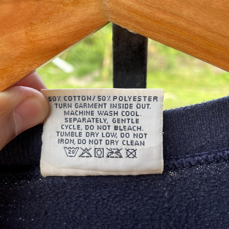May include: A close-up of a navy blue garment tag with white text. The tag reads: 50% cotton/50% polyester, turn garment inside out, machine wash cool, separately, gentle cycle, do not bleach, tumble dry low, do not iron, do not dry clean.
